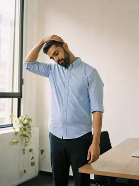 A man beside a desk, drawing his head down towards one shoulder