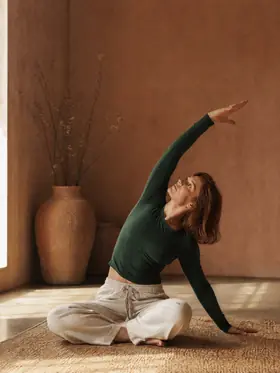 A woman sitting cross-legged, one arm sweeping overhead into a side bend